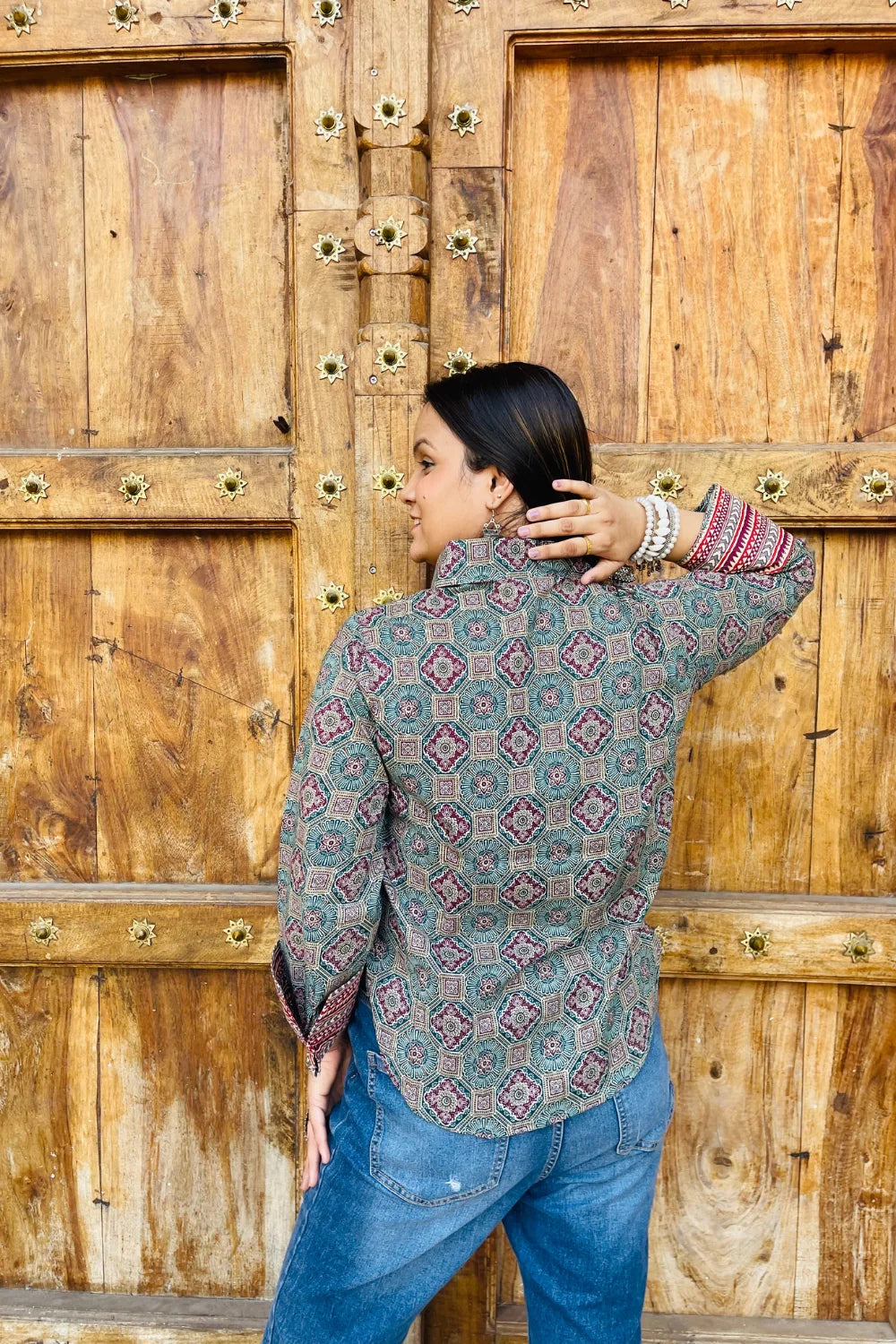 “Woman wearing the Sara cotton shirt with mixed geometric and stripe block prints in teal and red, relaxed fit, button‑down, standing before a carved wooden door”.