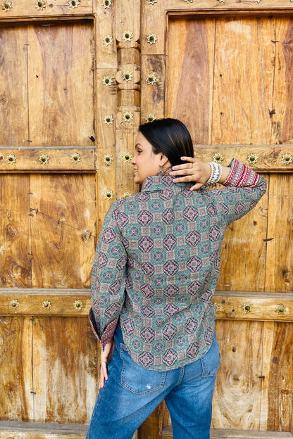 “Woman wearing the Sara cotton shirt with mixed geometric and stripe block prints in teal and red, relaxed fit, button‑down, standing before a carved wooden door”.
